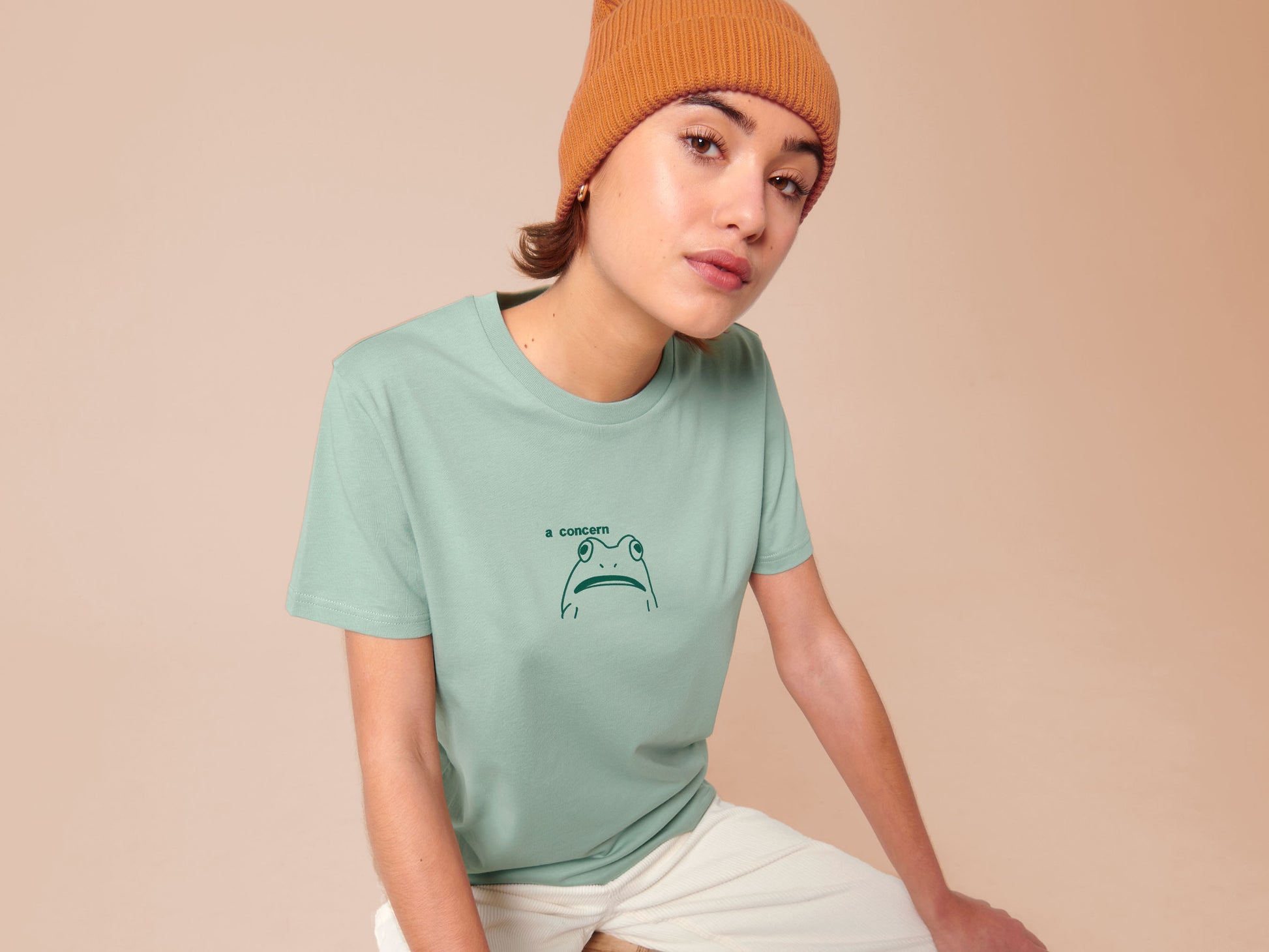 A woman sits wearing a green crew neck short sleeve t-shirt, with an embroidered green thread design of cute confused looking frog with the text a concern.