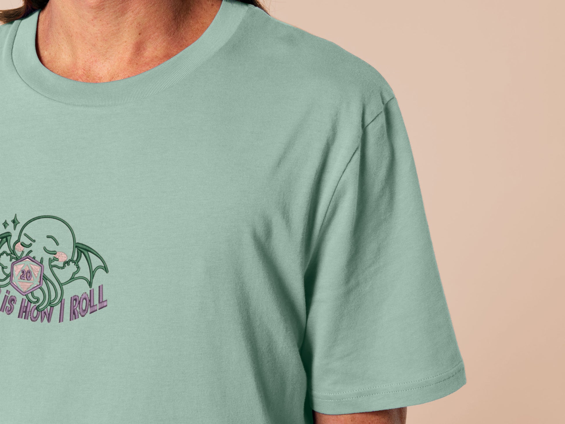 An embroidered green t-shirt design of a cute Cthullu with a D20 dice surrounded by the quote This Is How I Roll