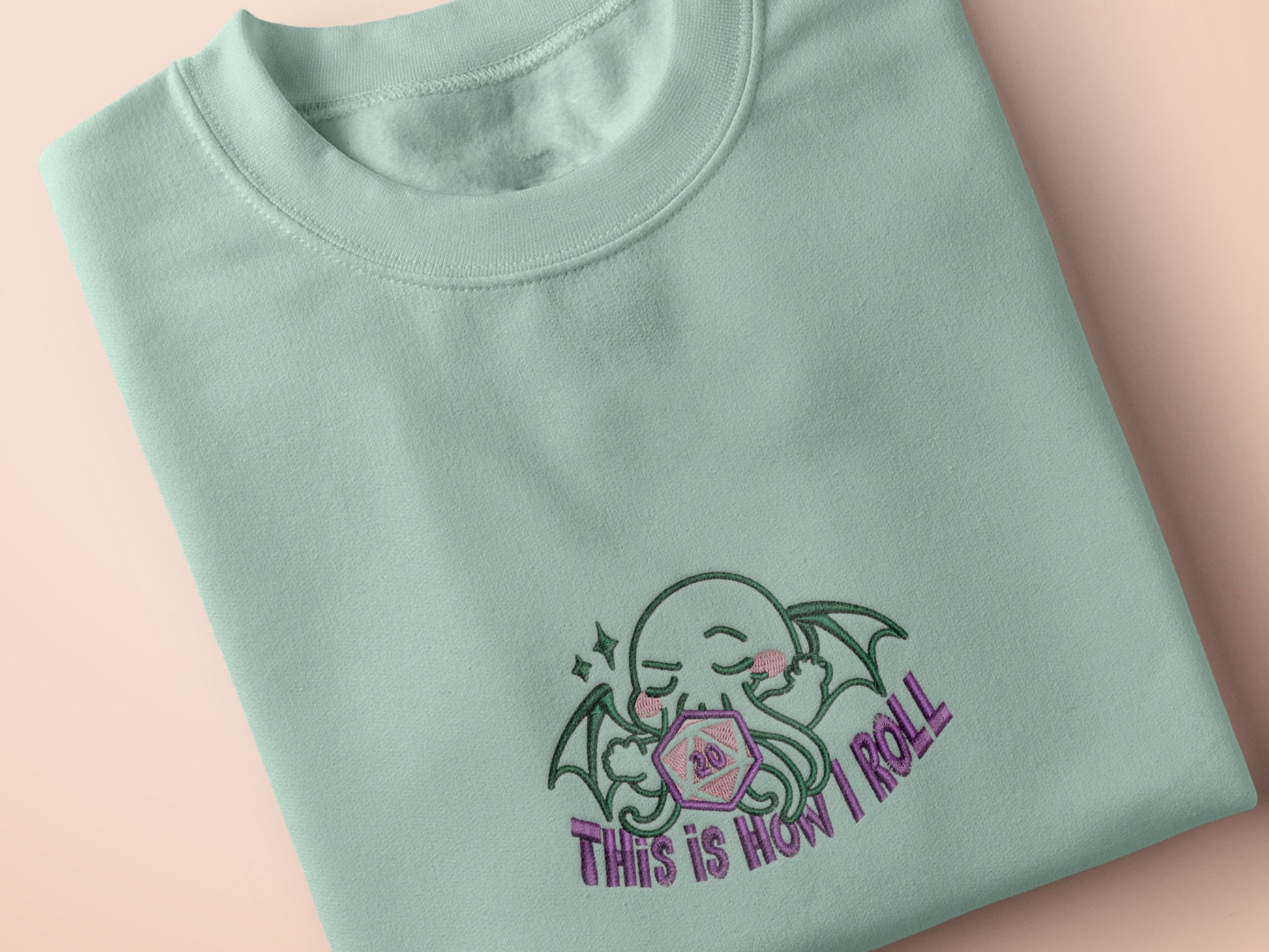 An embroidered green t-shirt design of a cute Cthullu with a D20 dice surrounded by the quote This Is How I Roll