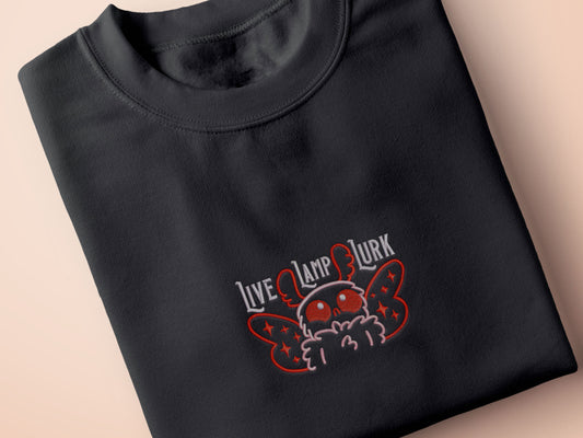 A dark grey crew neck short sleeve t-shirt, with an embroidered chibi mothman and the quote Live Lamp Lurk.