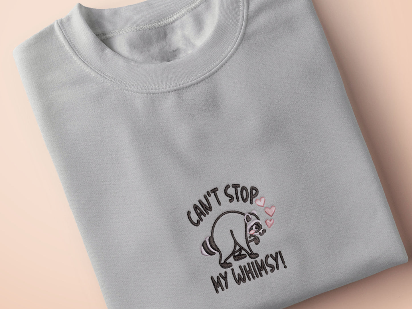 An Embroidered light grey t-shirt design of a cute Jimothy the viral disabled raccoon and the quote can't stop my whimsy!