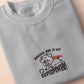 An Embroidered light grey t-shirt design of a cute chibsuper dog wearing a red cape panicked at a dice with the text rolling one is my superpower