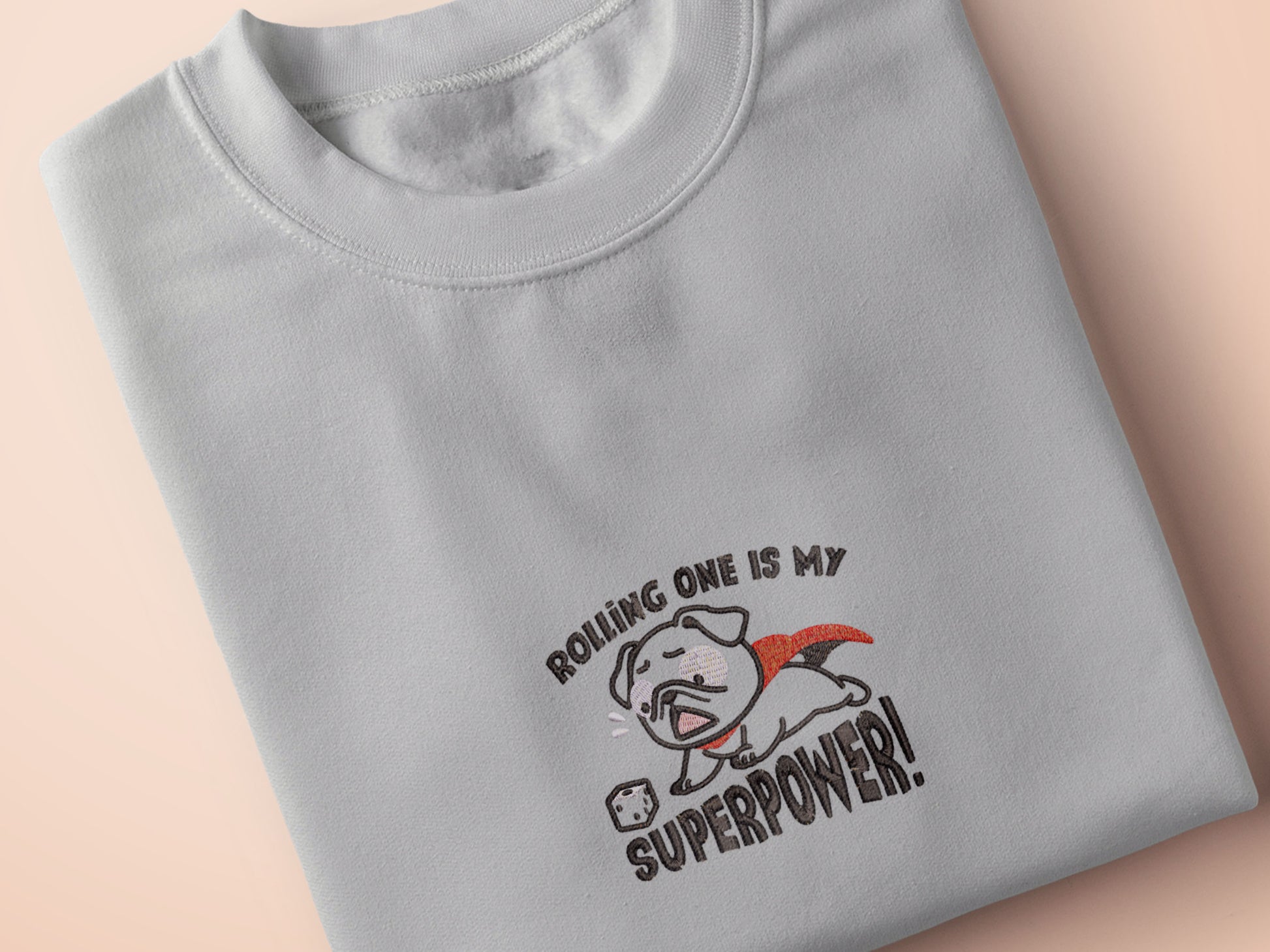 An Embroidered light grey t-shirt design of a cute chibsuper dog wearing a red cape panicked at a dice with the text rolling one is my superpower