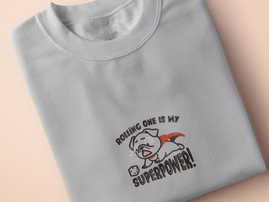 An Embroidered light grey t-shirt design of a cute chibsuper dog wearing a red cape panicked at a dice with the text rolling one is my superpower
