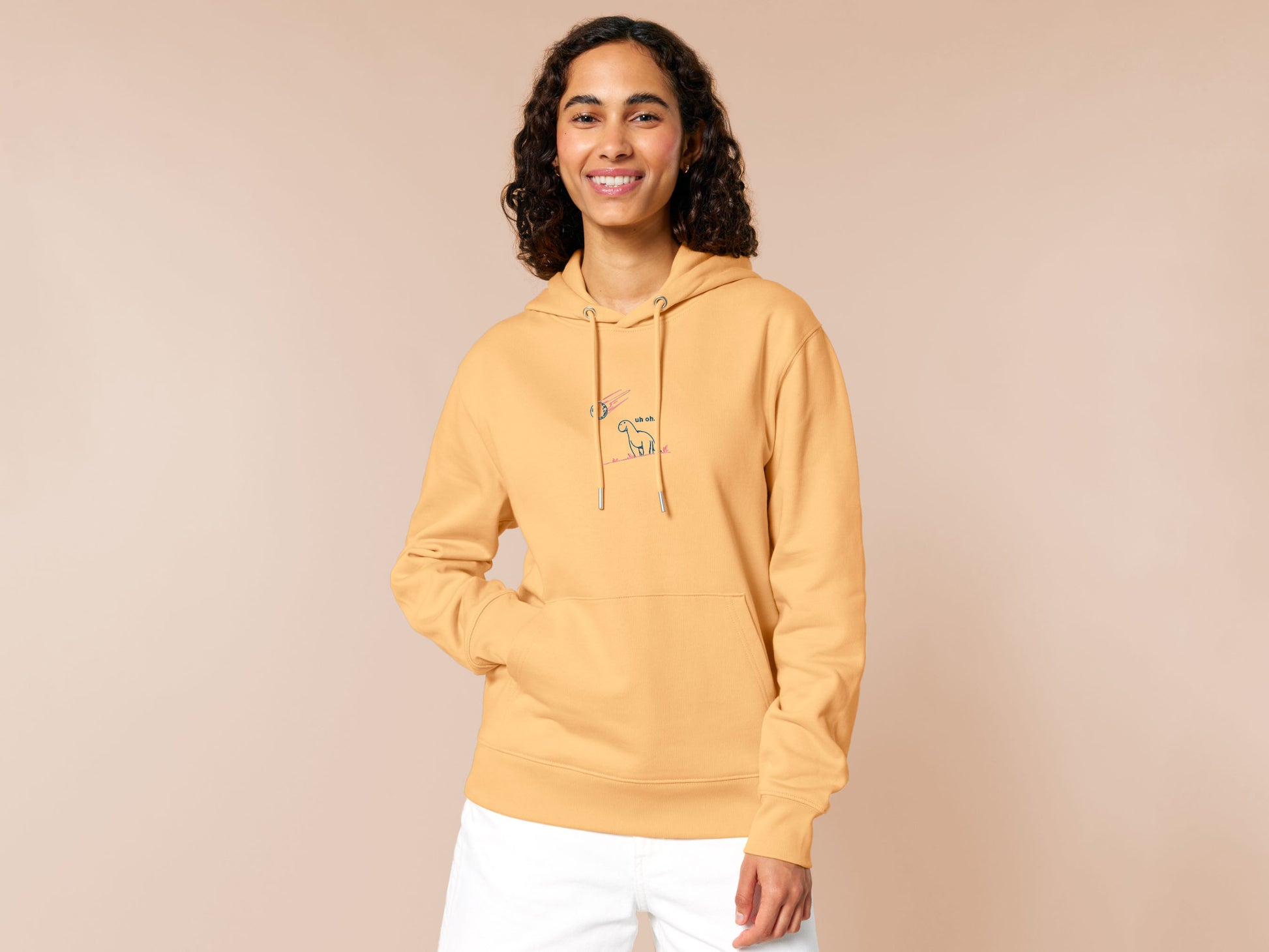 A yellow hoodie with a blue and pink embroidered design of a cute smiling dinosaur looking at a meteor plummeting to earth with the text uh oh...