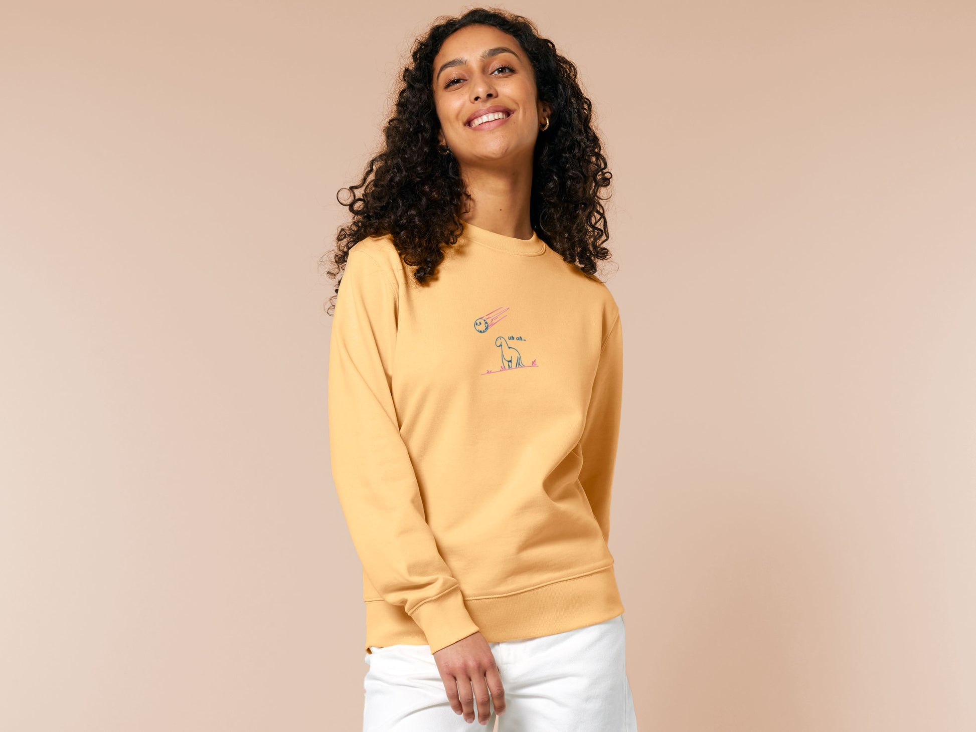 A yellow sweater with a blue and pink embroidered design of a cute smiling dinosaur looking at a meteor plummeting to earth with the text uh oh...