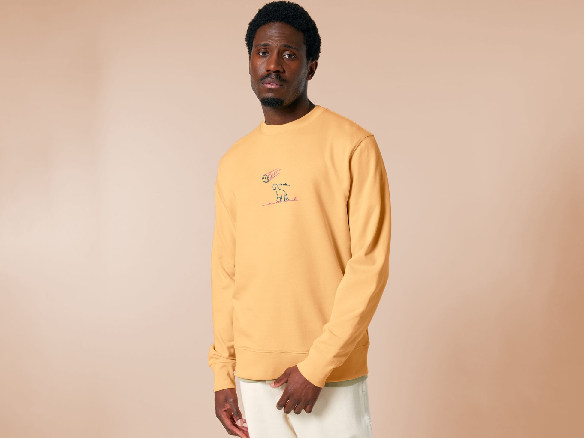 A yellow sweater with a blue and pink embroidered design of a cute smiling dinosaur looking at a meteor plummeting to earth with the text uh oh...