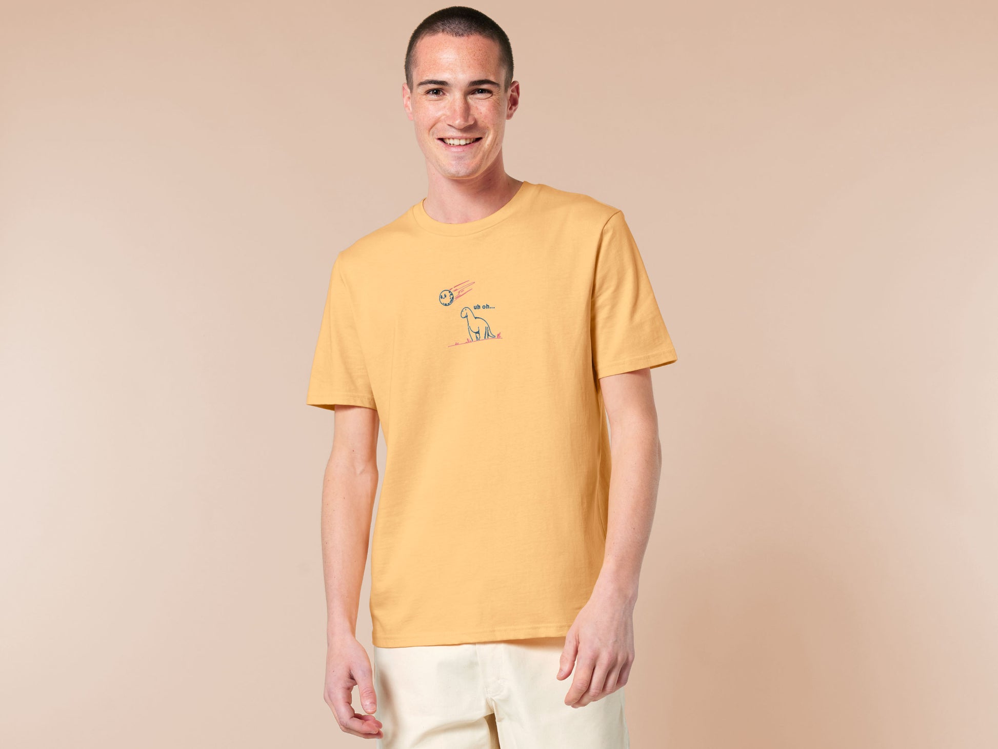 A yellow t-shirt with a blue and pink embroidered design of a cute smiling dinosaur looking at a meteor plummeting to earth with the text uh oh...