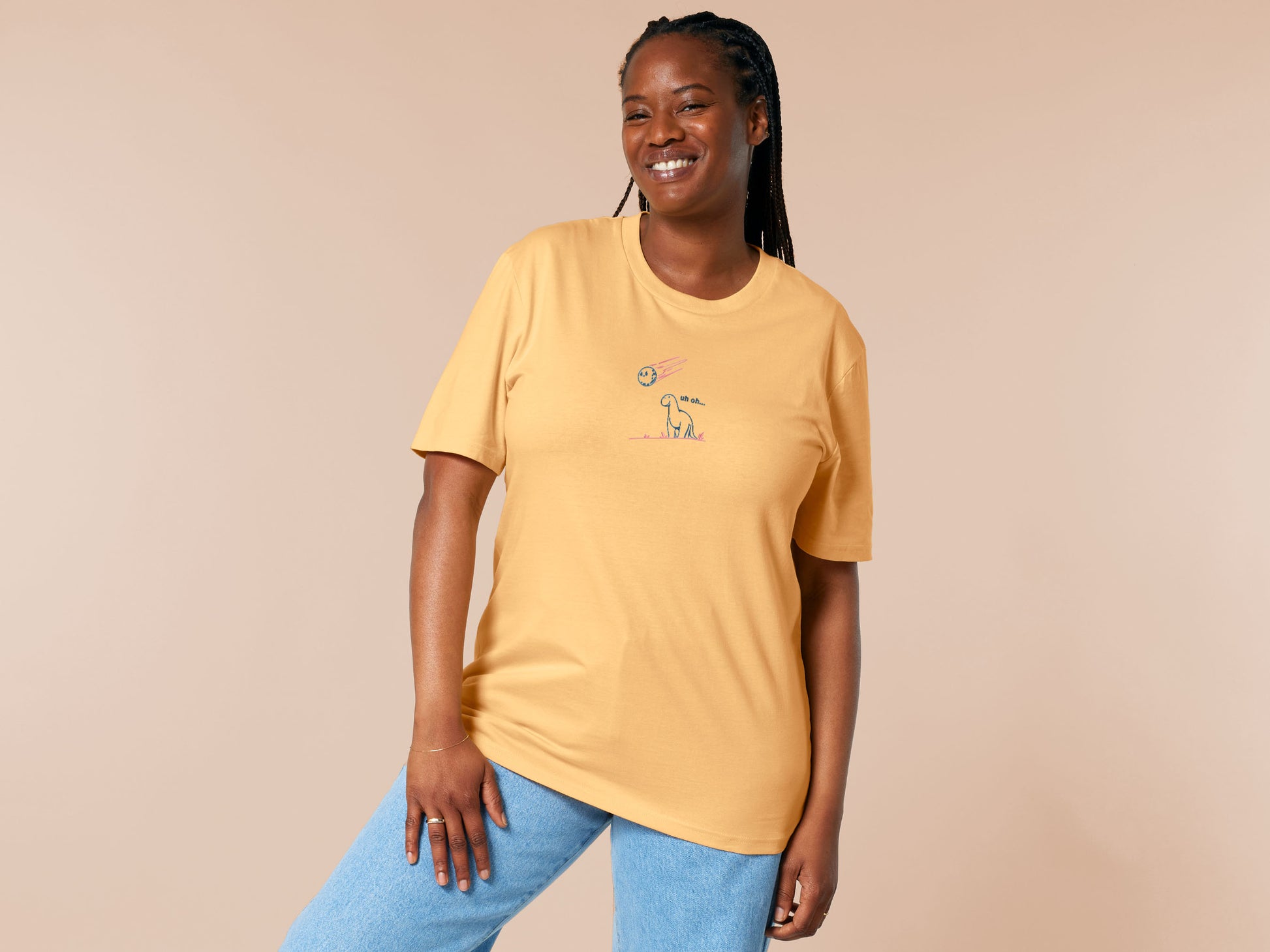 A yellow t-shirt with a blue and pink embroidered design of a cute smiling dinosaur looking at a meteor plummeting to earth with the text uh oh...