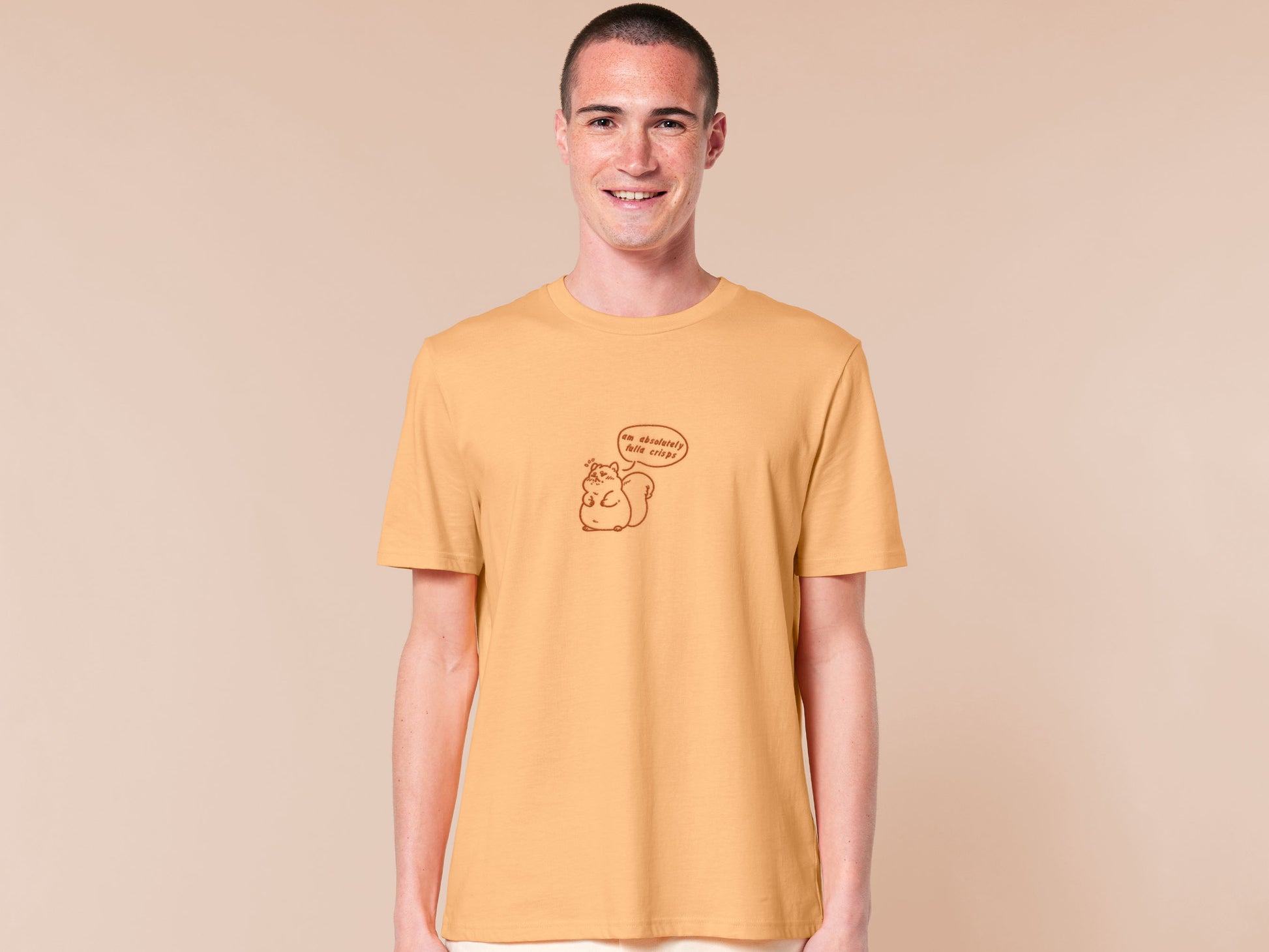 A man wearing a short sleeved yellow t-shirt with an embroidered design of a cute fat squirrel and a speech bubble with the text am absolutely fulla crisps