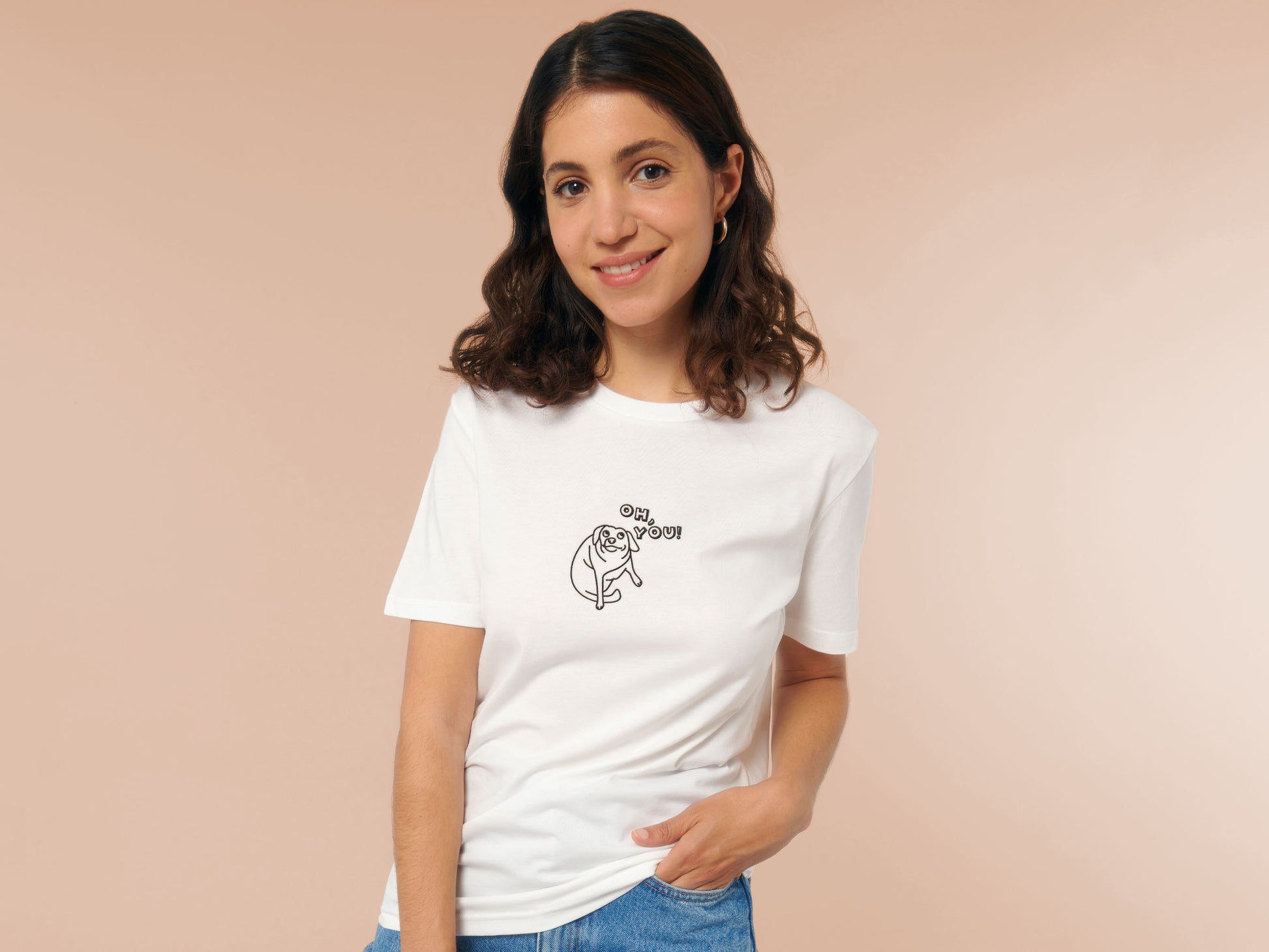 Woman wearing an off white crew neck short sleeve t-shirt, with an embroidered black thread design of cute meme dog with cheeky expression and big eyes, with the text Oh You!