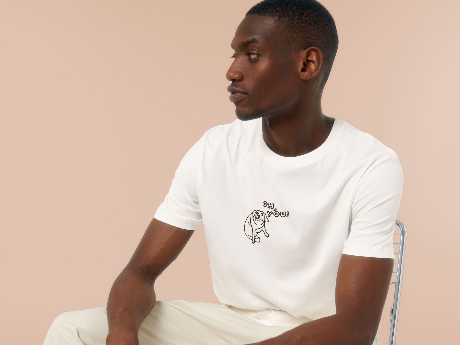 Man wearing an off white crew neck short sleeve t-shirt, with an embroidered black thread design of cute meme dog with cheeky expression and big eyes, with the text Oh You!