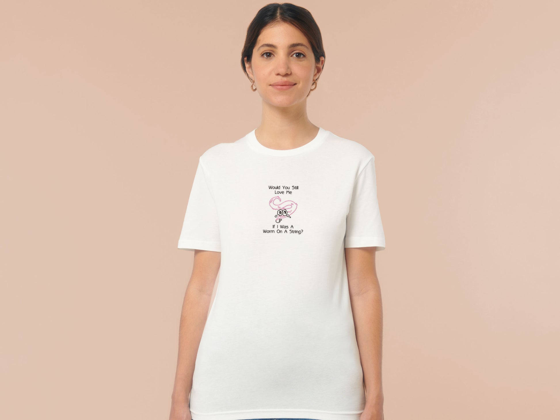A woman wearing an off white short sleeved crew neck t-shirt with an embroidered design of a pink worm on a string with a cute face crying with the black text Would You Still Love Me If I Was A Worm On A String