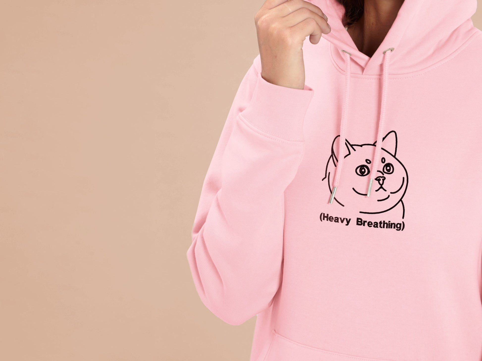 A woman wearing a pink long sleeve fleece hoodie, with an embroidered brown thread design of cute fat cat portrait with text underneath saying (Heavy Breathing)