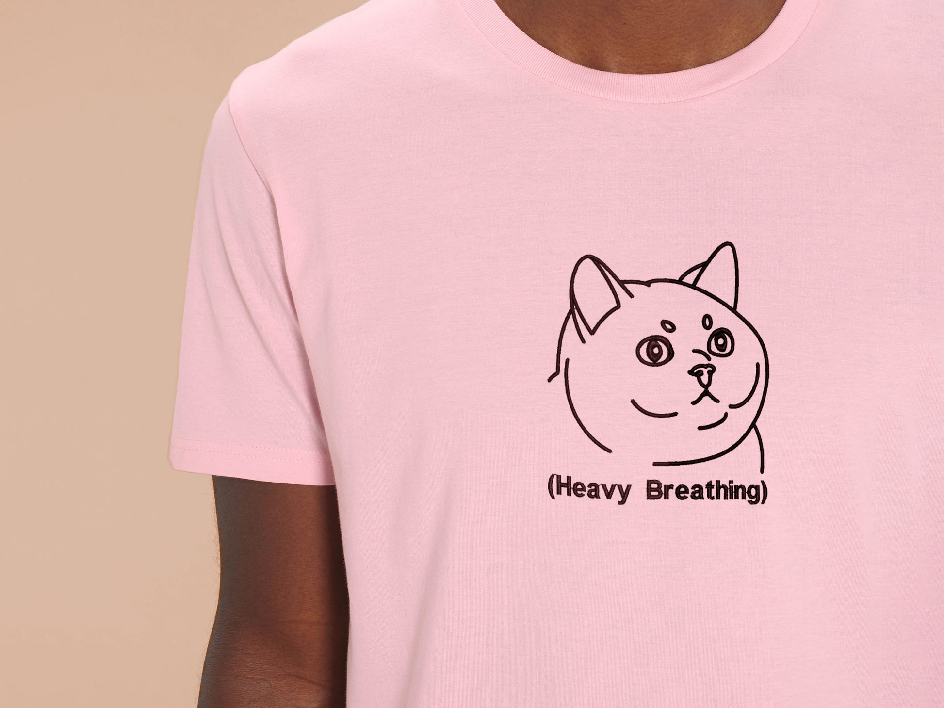 A man wearing a pink crew neck short sleeve t-shirt, with an embroidered brown thread design of cute fat cat portrait with text underneath saying (Heavy Breathing)