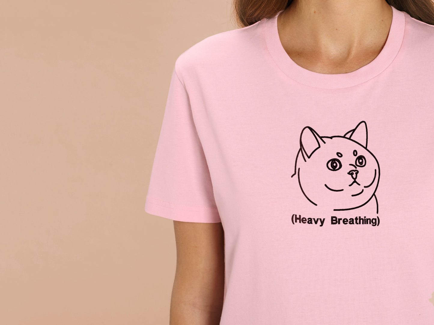 A woman wearing a pink crew neck short sleeve t-shirt, with an embroidered brown thread design of cute fat cat portrait with text underneath saying (Heavy Breathing)