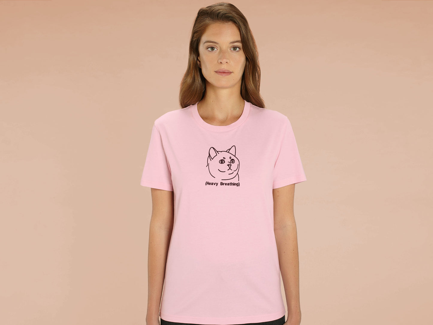 A woman wearing a pink crew neck short sleeve t-shirt, with an embroidered brown thread design of cute fat cat portrait with text underneath saying (Heavy Breathing)