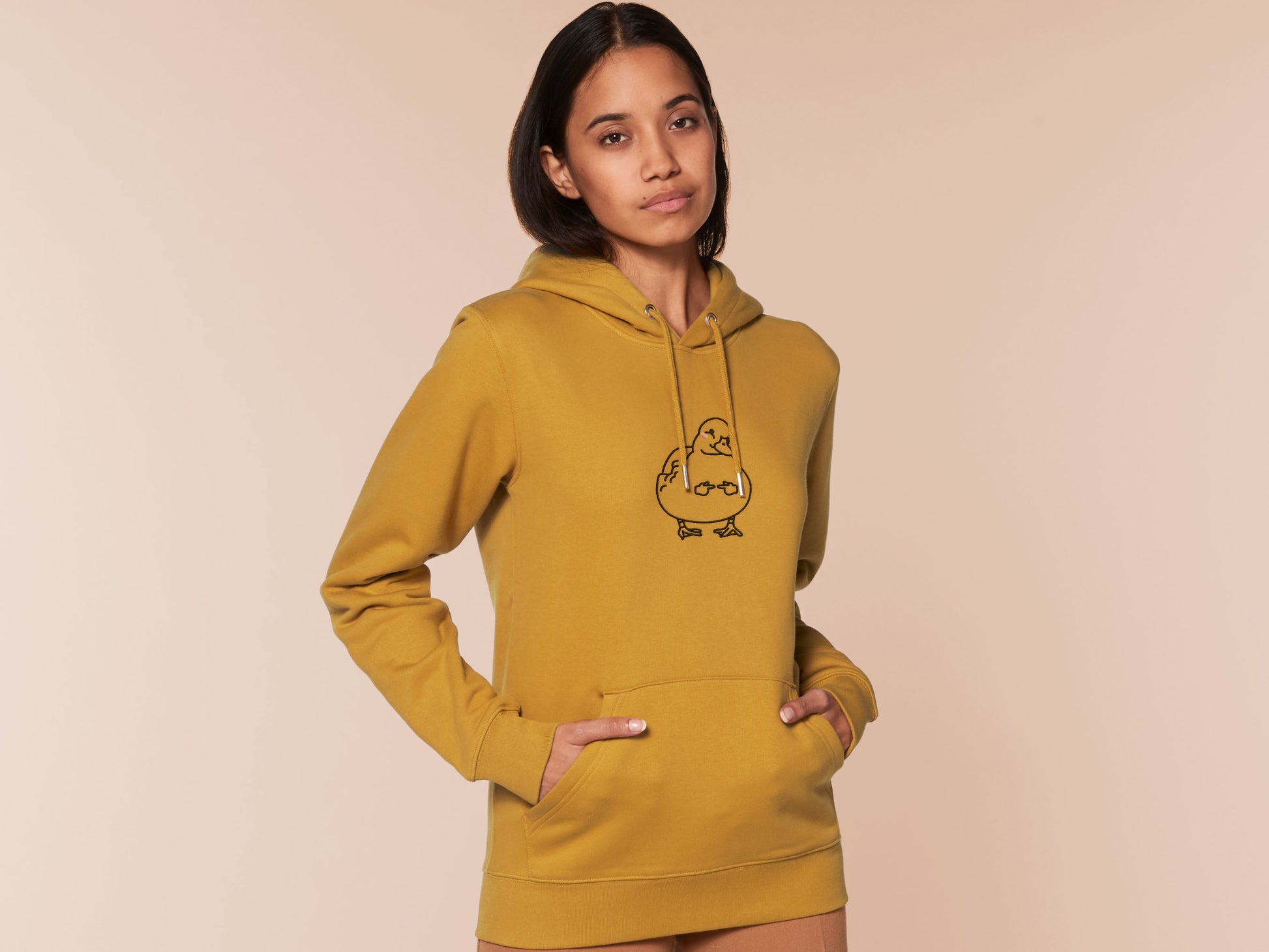 A woman wearing a yellow long sleeve fleece hoodie, with an embroidered black thread design of cute blushing duck with the for me finger hand emoji symbols