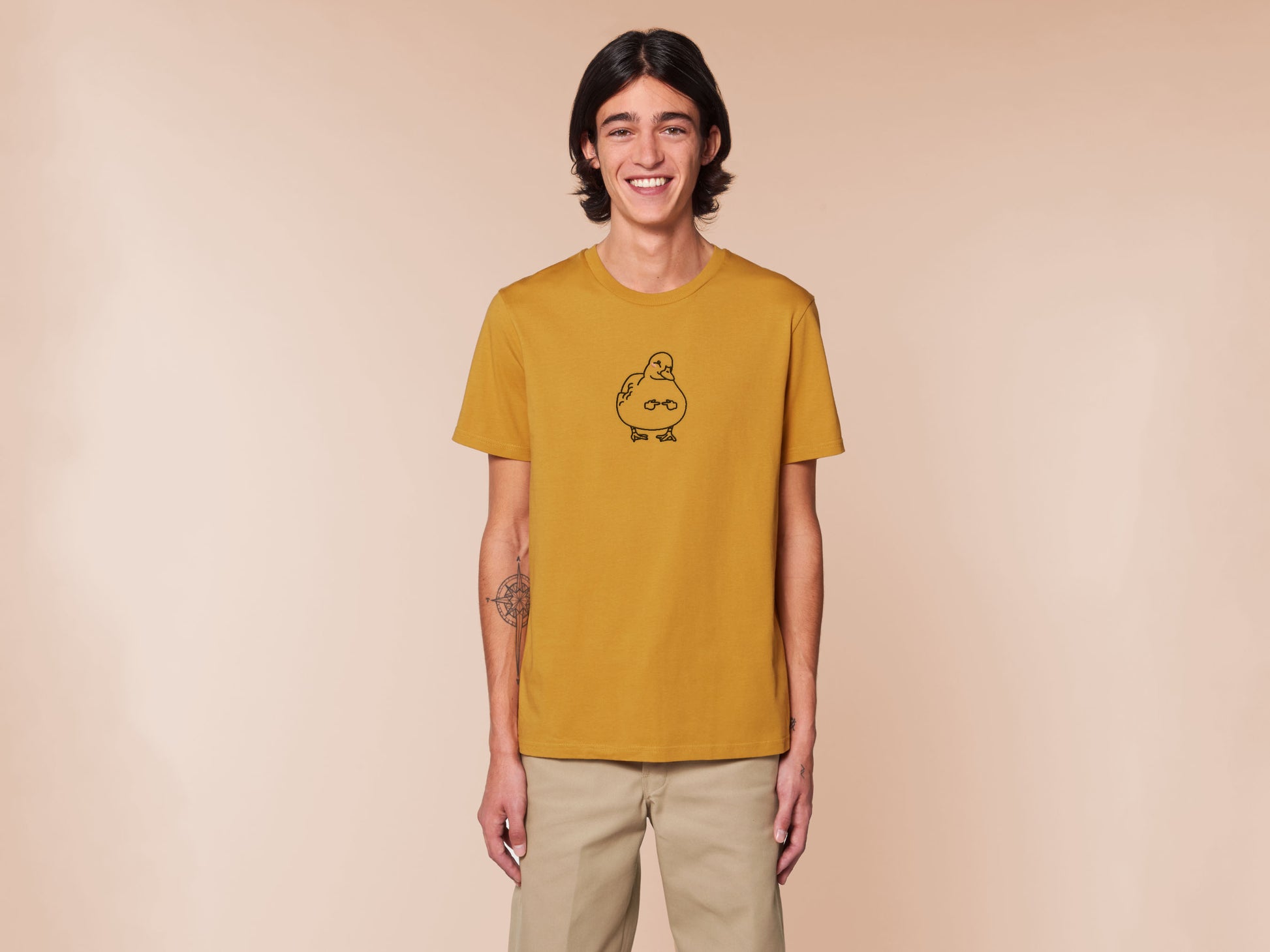 A male wearing a yellow crew neck short sleeve t-shirt, with an embroidered black thread design of cute blushing duck with the for me finger hand emoji symbols