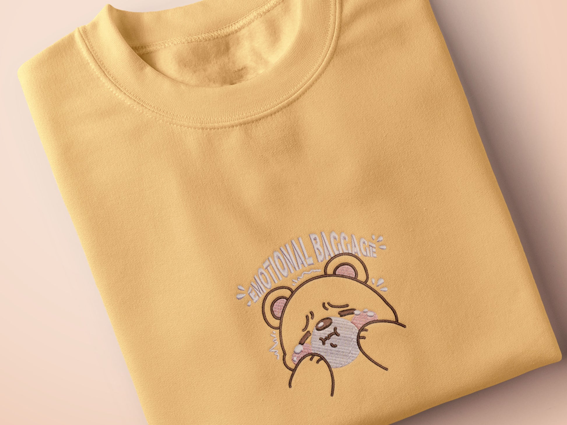 A yellow crew neck short sleeve t-shirt, with an embroidered brown thread design of a cute panicking bear and the quote emotional baggage