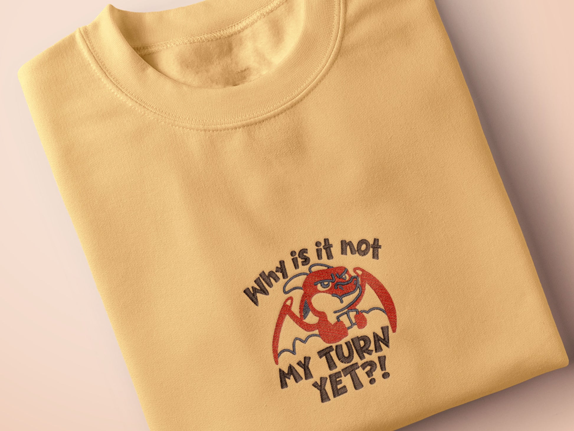 A yellow tshirt with an embroidered design of a cute grumpy red dragon playing a card game with the text why is it not my turn yet?!