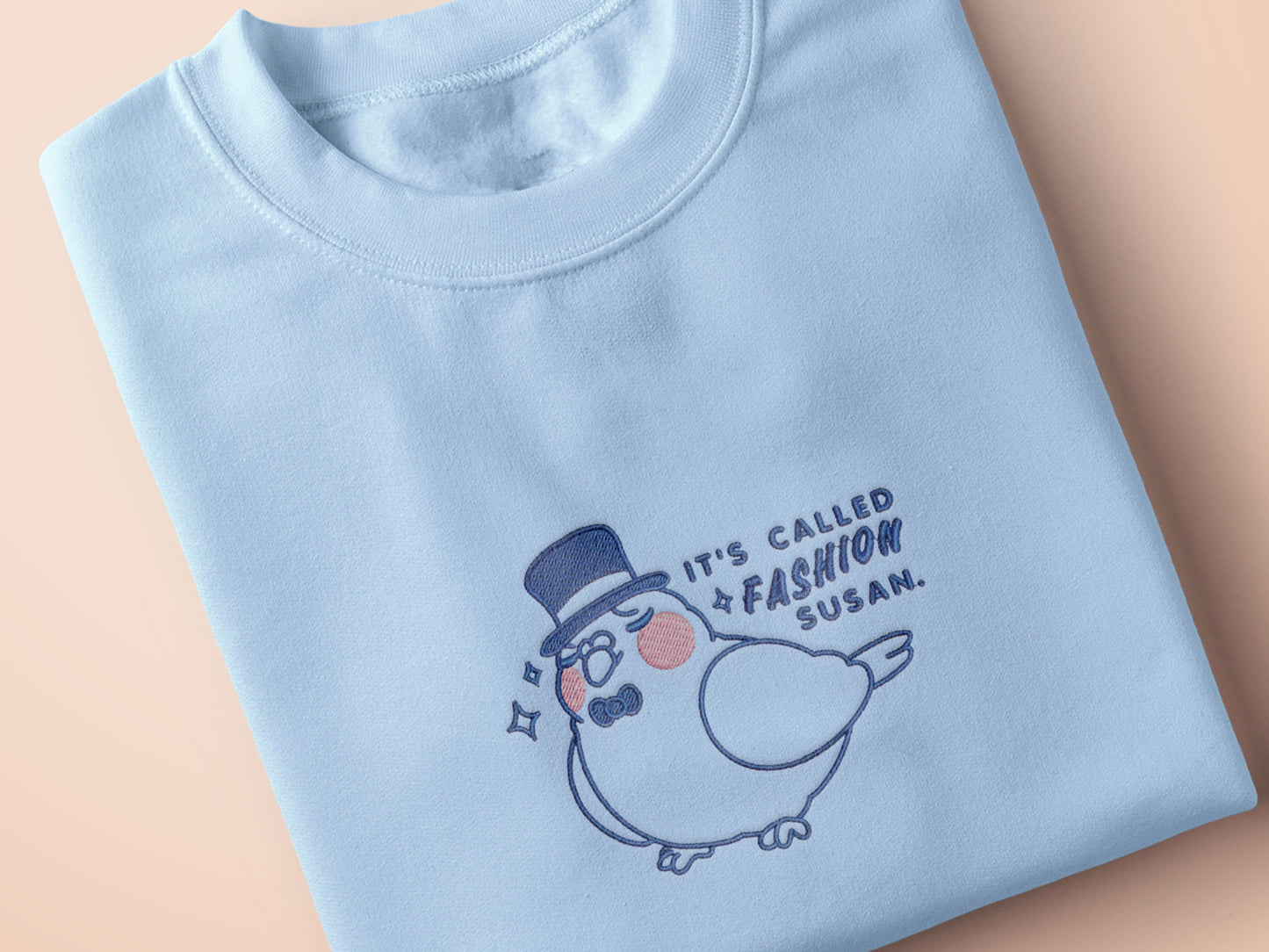 An embroidered blue t-shirt design of a cute pigeon wearing a top hat and the text It's Called Fashion Susan