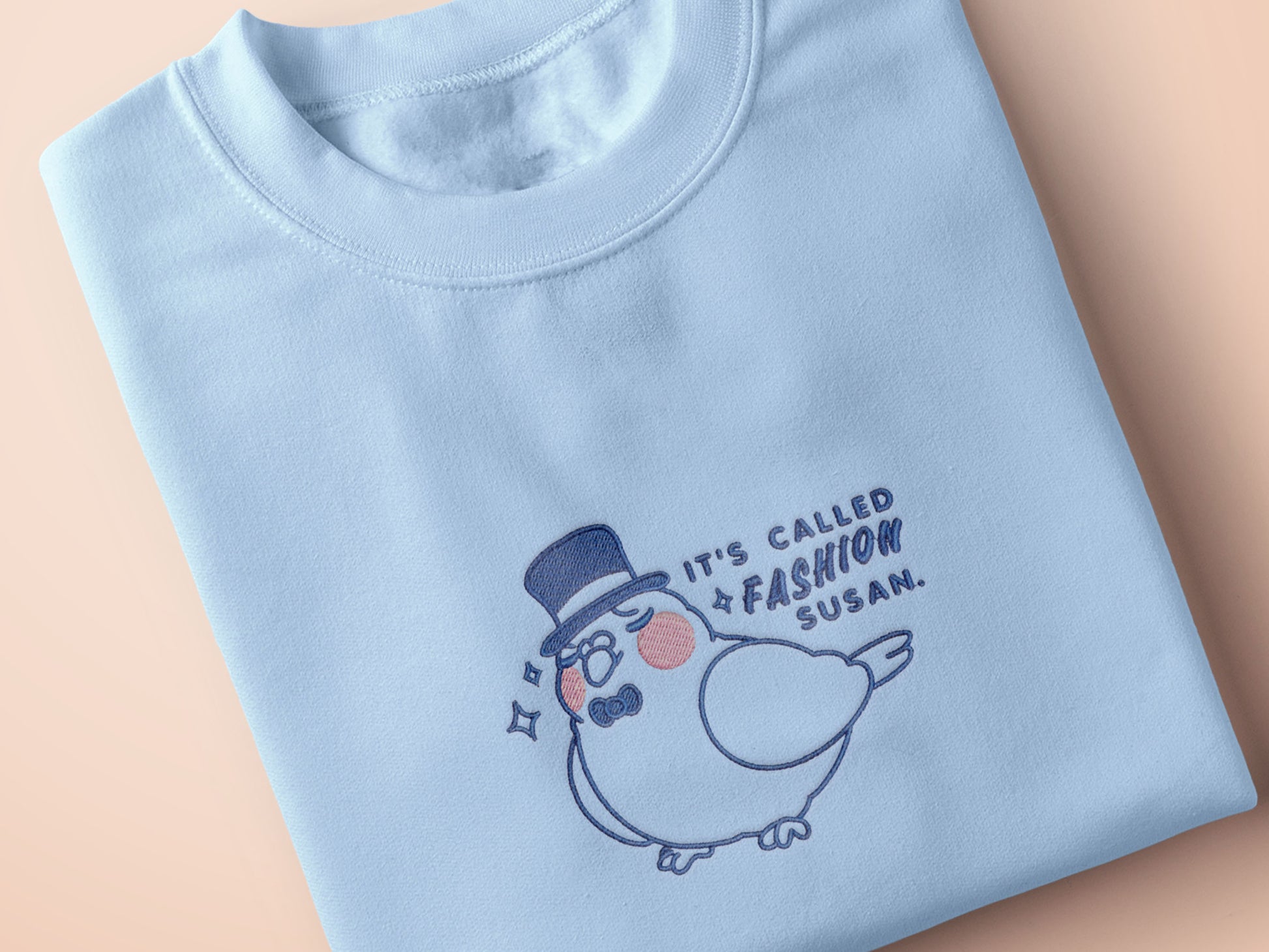 An embroidered blue t-shirt design of a cute pigeon wearing a top hat and the text It's Called Fashion Susan