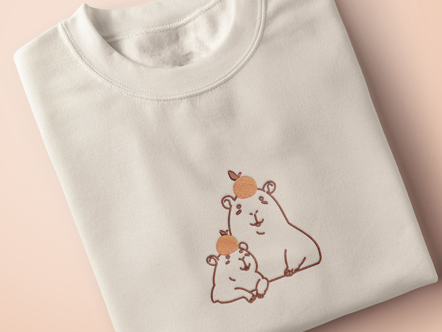 Embroidered cream t-shirt design of two cute chibi capybaras with oranges on their heads.