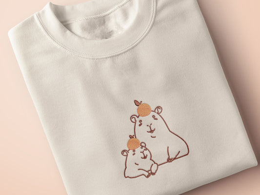 Embroidered cream t-shirt design of two cute chibi capybaras with oranges on their heads.