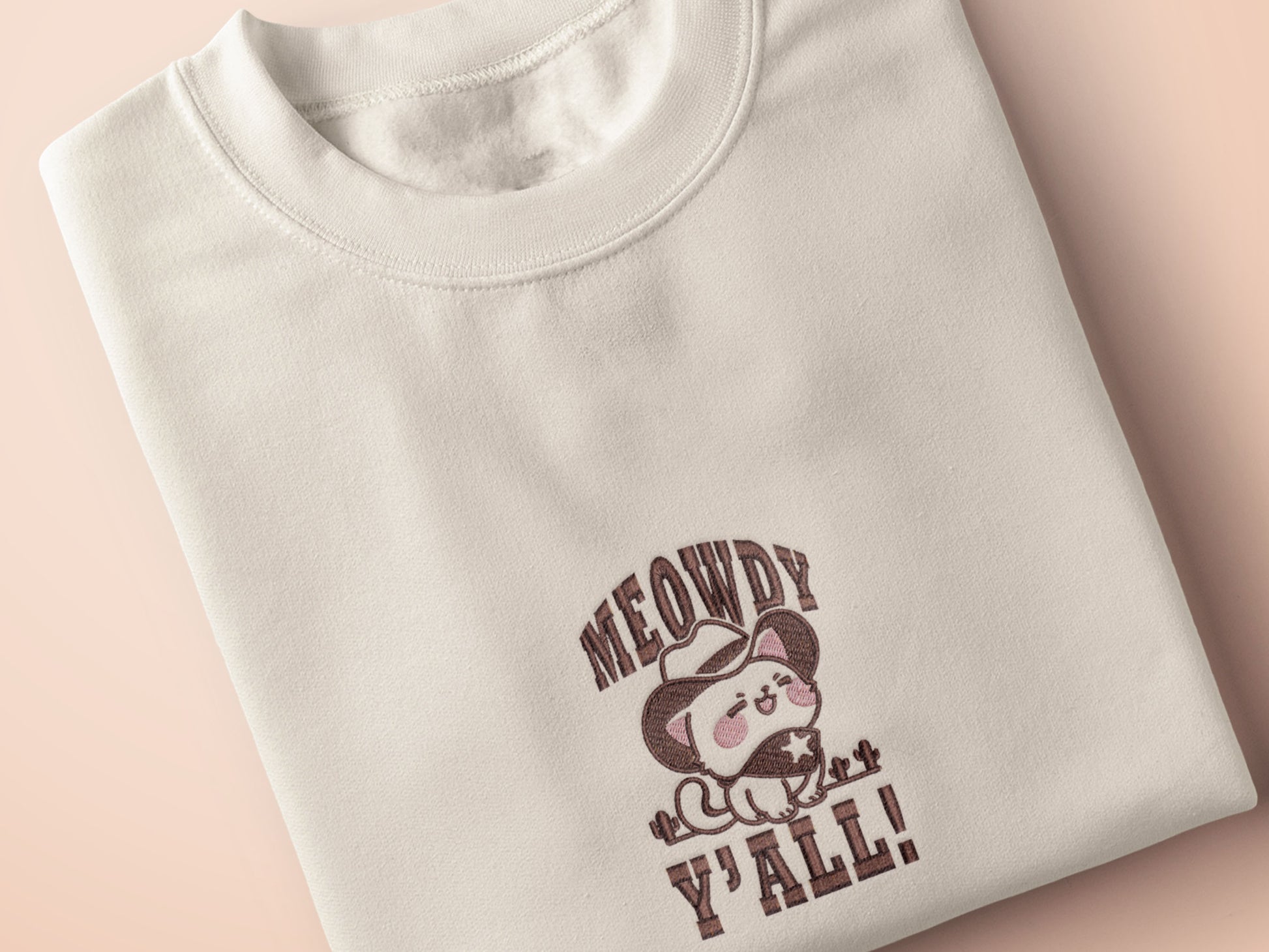 A cream crew neck short sleeve t-shirt, with an embroidered brown thread design of a cute cowboy cat with hat surrounded by the quote meowfy ya'll!