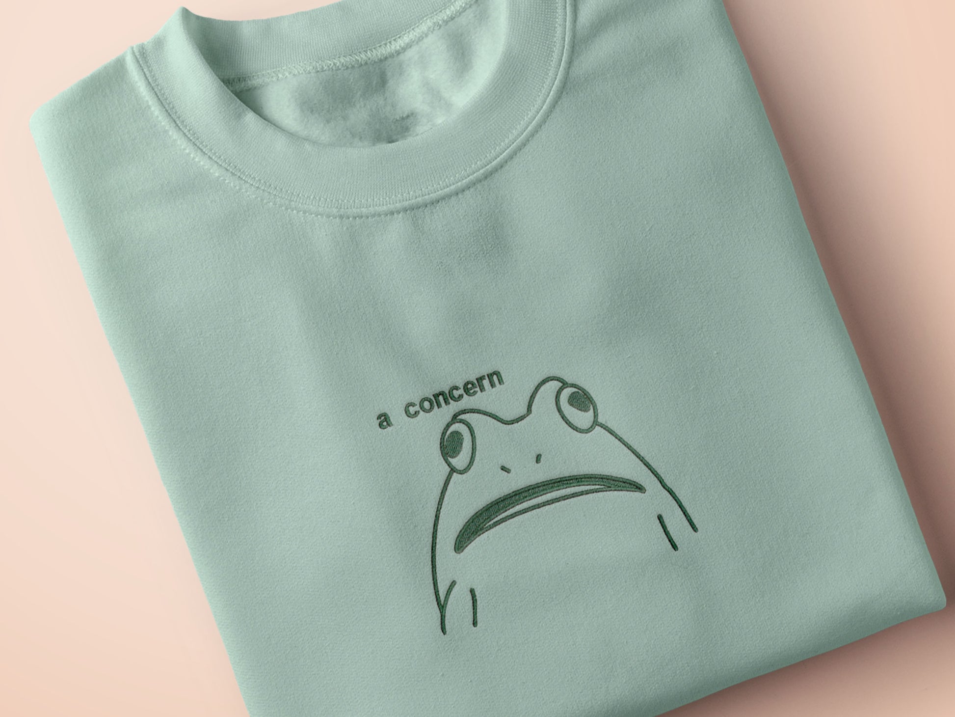 An embroidered green t-shirt design of a funny cartoon frog with the text a concern.