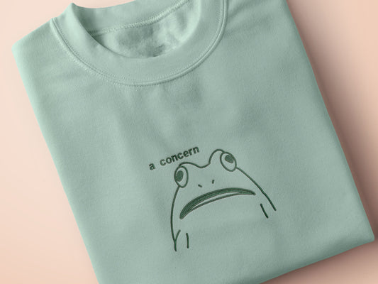 An embroidered green t-shirt design of a funny cartoon frog with the text a concern.