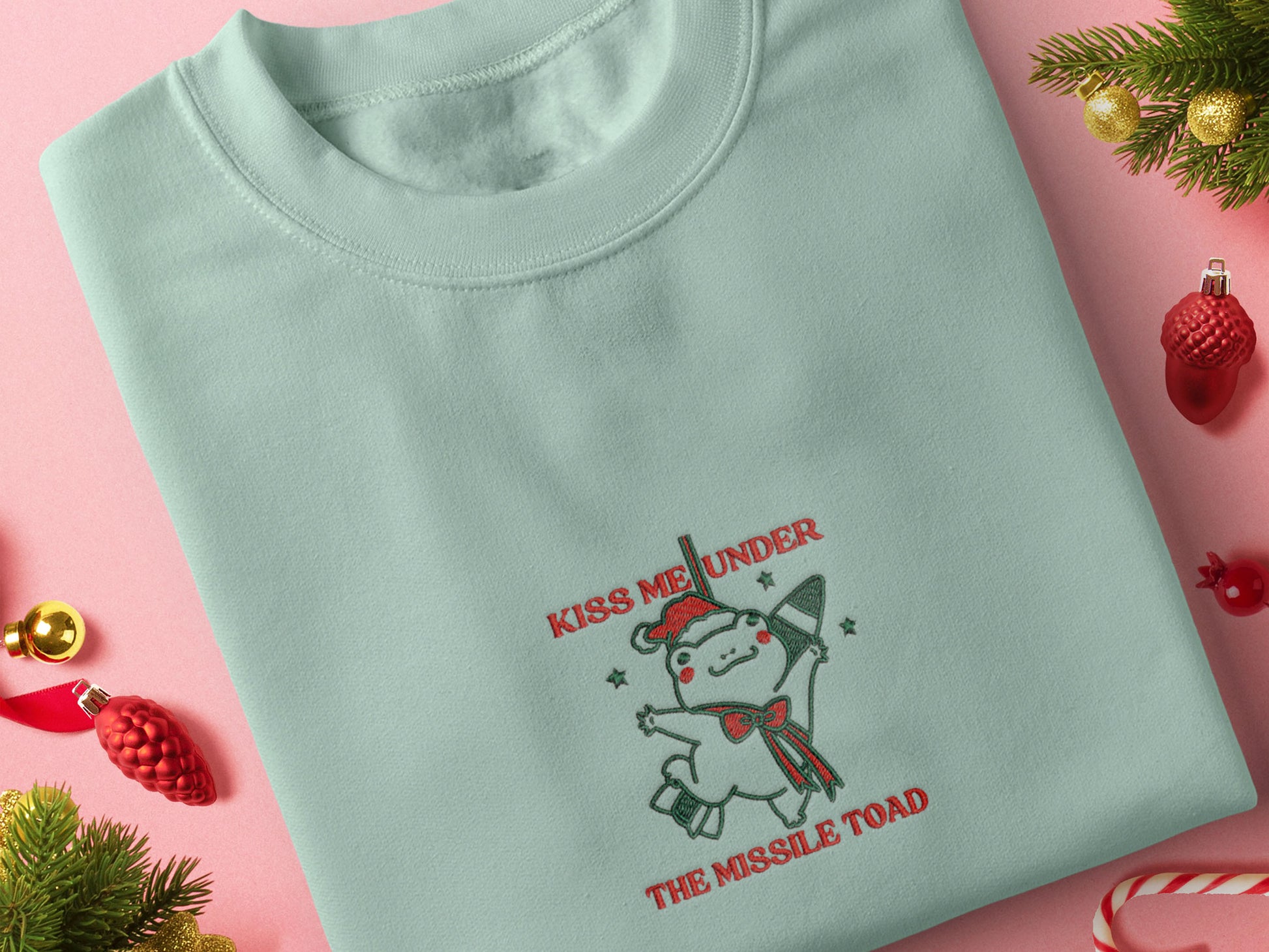 Green Christmas jumper with an embroidered design of the viral frog
meme Missile Toad, featuring a cute hanging chibi Christmas frog with the quote kiss me under the missile toad