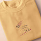 A yellow top with a blue and pink embroidered design of a cute smiling dinosaur looking at a meteor plummeting to earth with the text uh oh...
