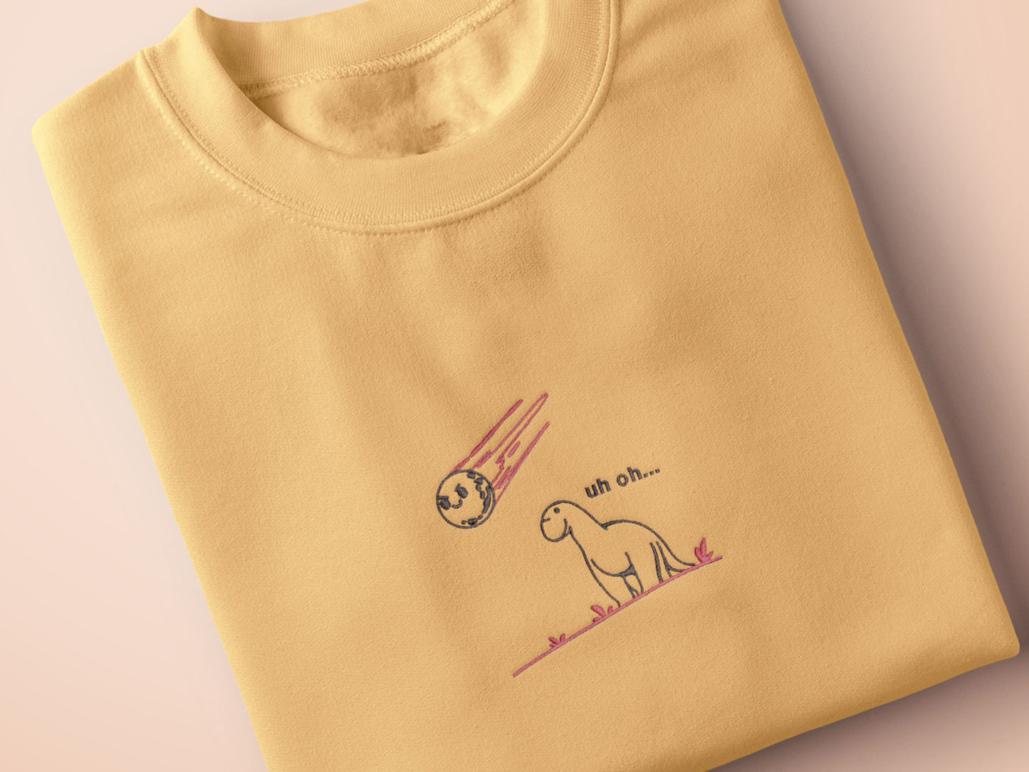 A yellow top with a blue and pink embroidered design of a cute smiling dinosaur looking at a meteor plummeting to earth with the text uh oh...