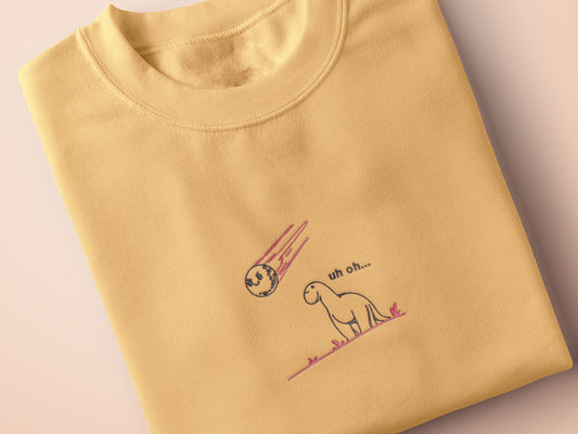 A yellow top with a blue and pink embroidered design of a cute smiling dinosaur looking at a meteor plummeting to earth with the text uh oh...