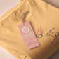 A yellow top with a blue and pink embroidered design of a cute smiling dinosaur looking at a meteor plummeting to earth with the text uh oh...