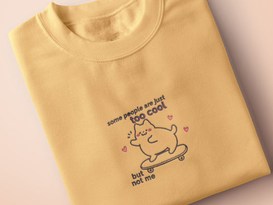Light yellow short sleeve t-shirt with embroidered pink and blue design of a cute chibi skateboarding cat with the text some people are just too cool but not me.