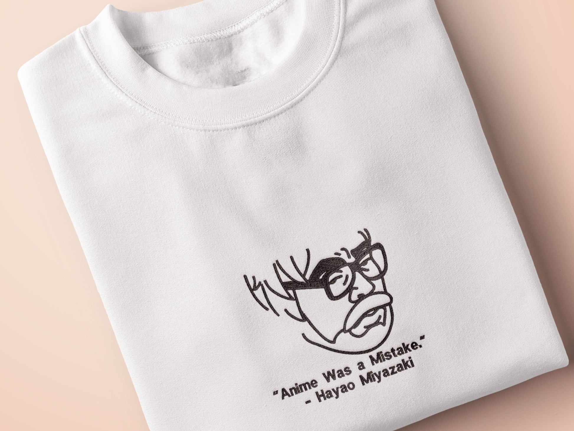 A off white crew neck short sleeve t-shirt, with an embroidered black thread design of a portrait of a depressed Hayao Miyazaki the creator of Studio Ghibli with text underneath that reads "Anime Was A Mistake." - Hayao Miyazaki