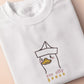 An embroidered white t-shirt design of a cute cartoon goose wearing a paper hat with text underneath that reads you silly goose.