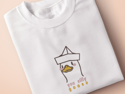 An embroidered white t-shirt design of a cute cartoon goose wearing a paper hat with text underneath that reads you silly goose.