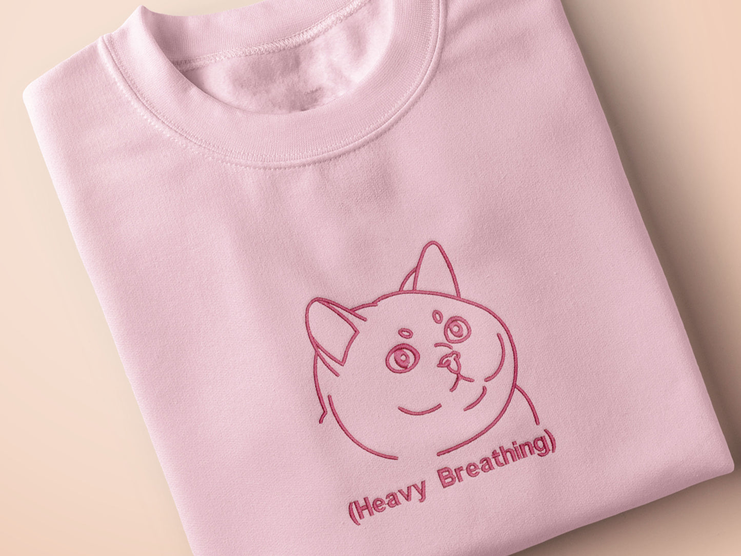 A pink crew neck short sleeve t-shirt, with an embroidered pink thread design of a cute fat cat portrait with text underneath saying (Heavy Breathing)