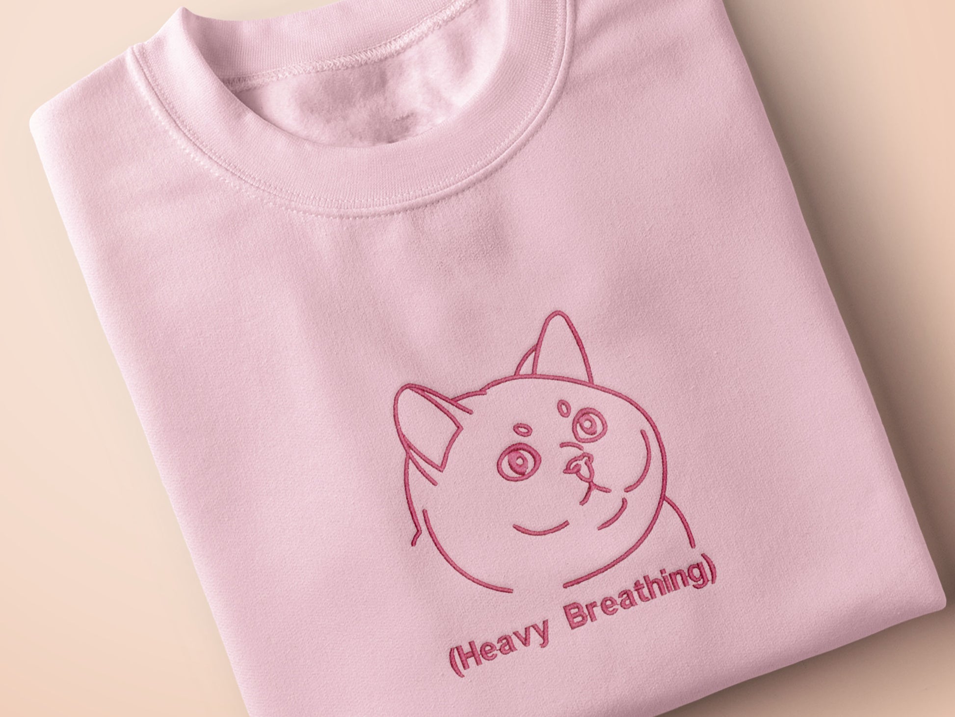 A pink crew neck short sleeve t-shirt, with an embroidered pink thread design of a cute fat cat portrait with text underneath saying (Heavy Breathing)