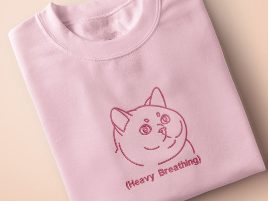 A pink crew neck short sleeve t-shirt, with an embroidered pink thread design of a cute fat cat portrait with text underneath saying (Heavy Breathing)