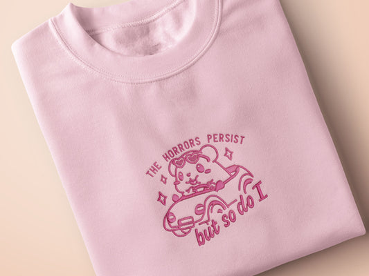 Pink embroidered t-shirt with a cute chibi guinea pig hamster wearing heart-shaped sunglasses and driving a little pink car surrounded by the quote the horrors persist but so do I.