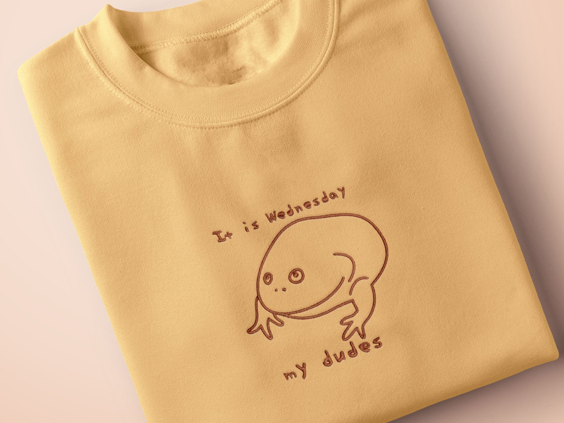 A yellow crew neck short sleeve t-shirt, with an embroidered black thread design of the Reddit meme rain frog with the text It is Wednesday my dudes