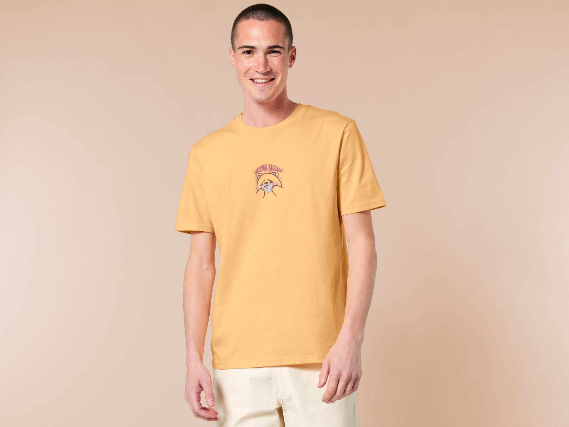 A yellow crew neck short sleeve t-shirt, with an embroidered brown thread design of a cute panicking bear and the quote emotional baggage