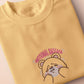 A yellow crew neck short sleeve t-shirt, with an embroidered brown thread design of a cute panicking bear and the quote emotional baggage