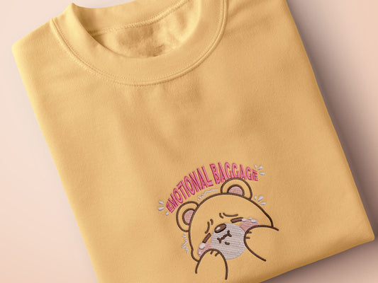A yellow crew neck short sleeve t-shirt, with an embroidered brown thread design of a cute panicking bear and the quote emotional baggage
