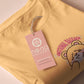A yellow crew neck short sleeve t-shirt, with an embroidered brown thread design of a cute panicking bear and the quote emotional baggage